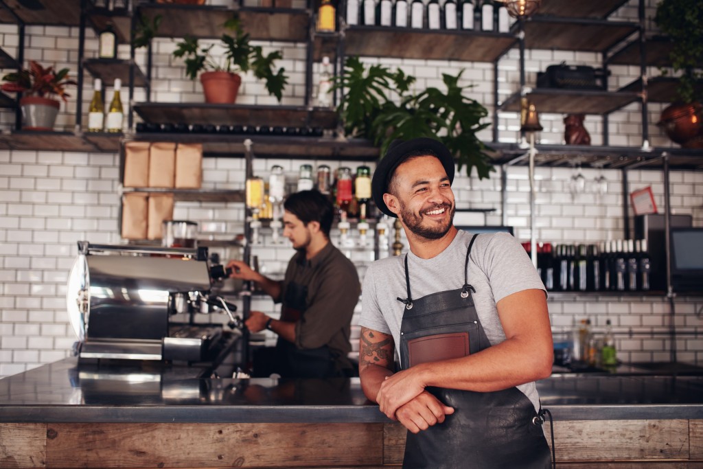 Portrait of cafe owner wearing a hat and apron standing at the counter and looking away.