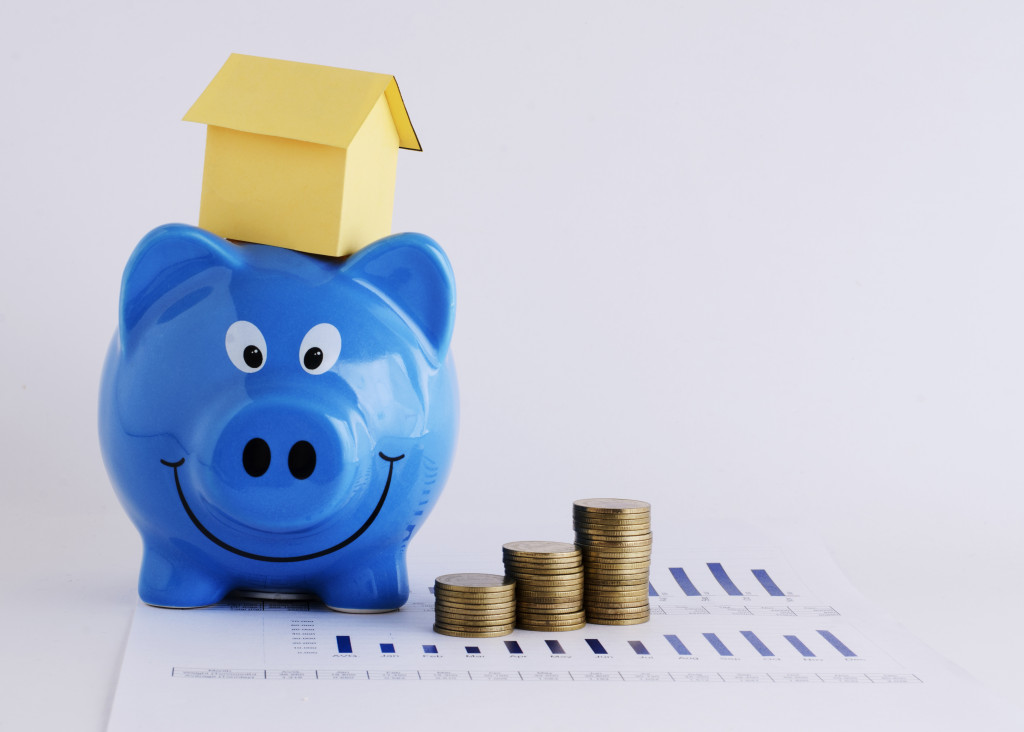 Blue piggy bank with a miniature house on top and coin stacks beside it